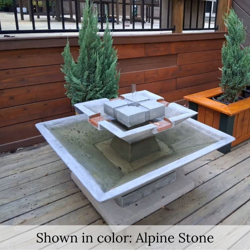 Facet Fountain with bubbling water cascading into a square basin on a wood deck surrounded by lush green plants in sideview.