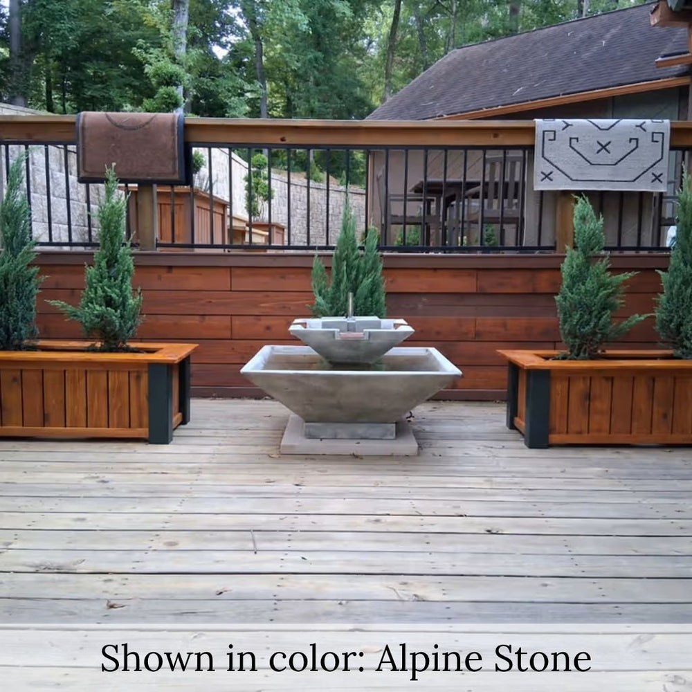 Facet Fountain with bubbling water cascading into a square basin on a wood deck surrounded by lush green plants.